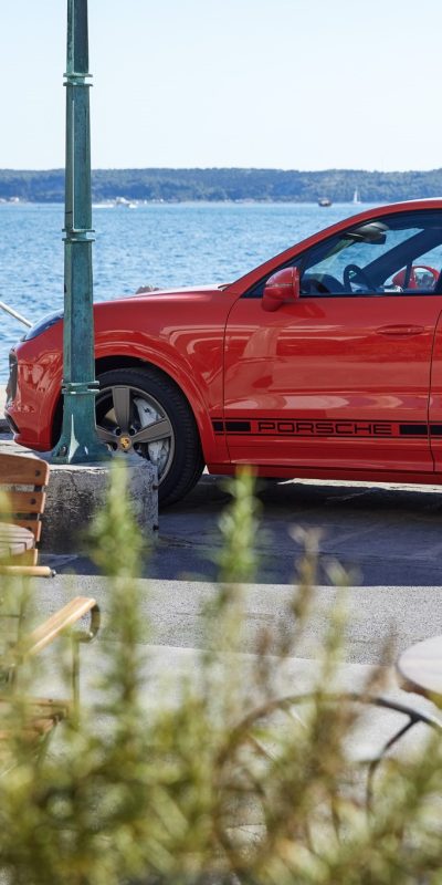 Punainen Porsche Cayenne kesäisissä maisemissa rannalla.